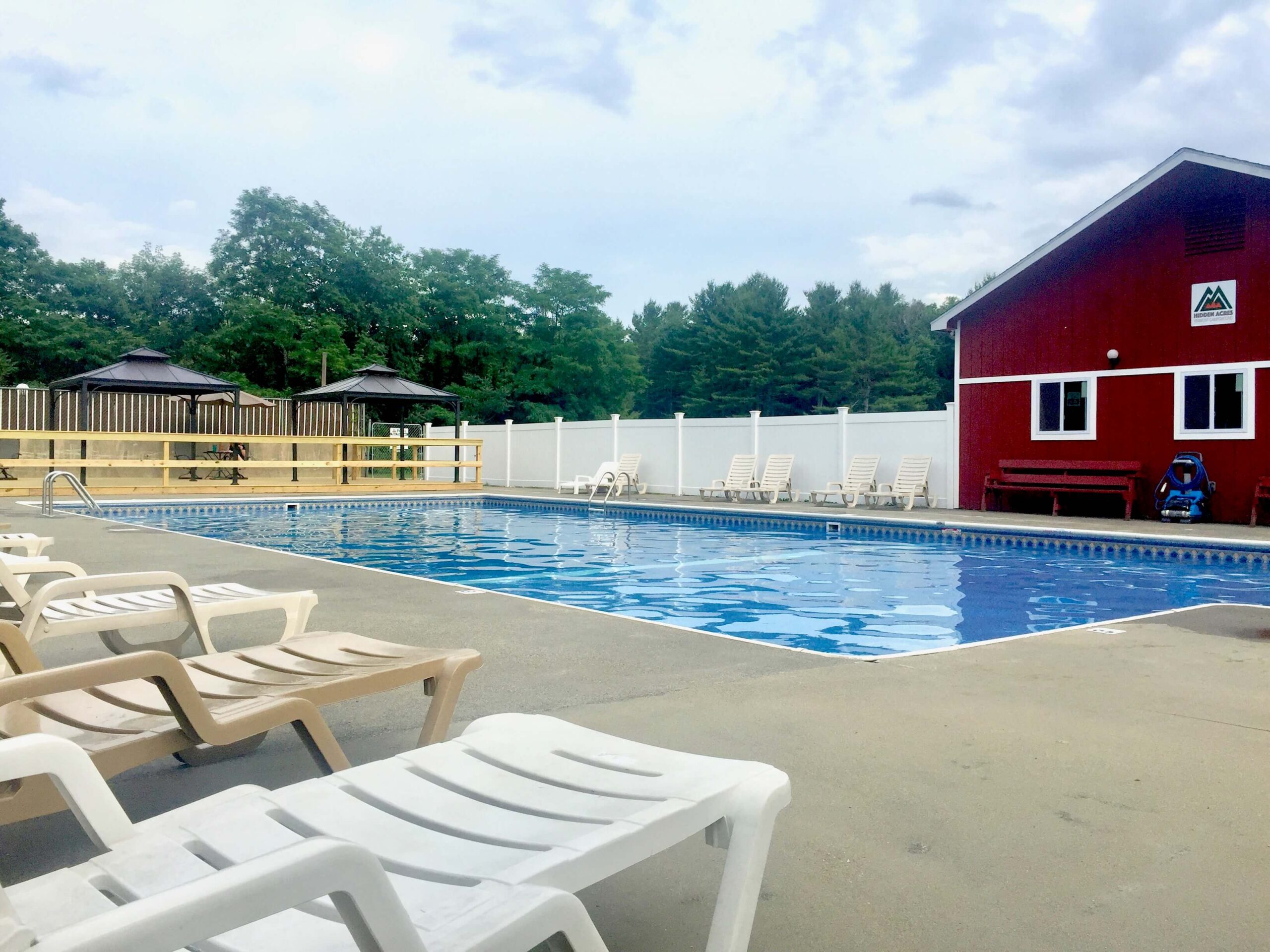 Outdoor swimming pool surrounded by white lounge chairs on concrete. A white fence borders the pool, with green trees in the background. A red building with windows is on the right. The sky is partly cloudy, creating a calm, relaxed atmosphere.