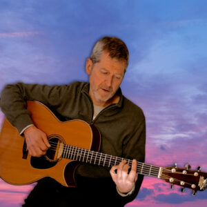 A man with short gray hair and a beard plays an acoustic guitar, looking down at his fingers on the fretboard. He wears a gray zip-up sweater. The background features a vibrant sky with blue, purple, and pink clouds at sunset.